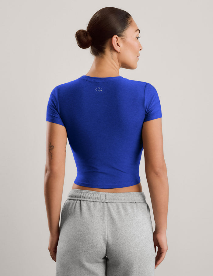 A woman modeling a royal blue short-sleeve crop top paired with light gray sweatpants, standing against a neutral light gray studio background.