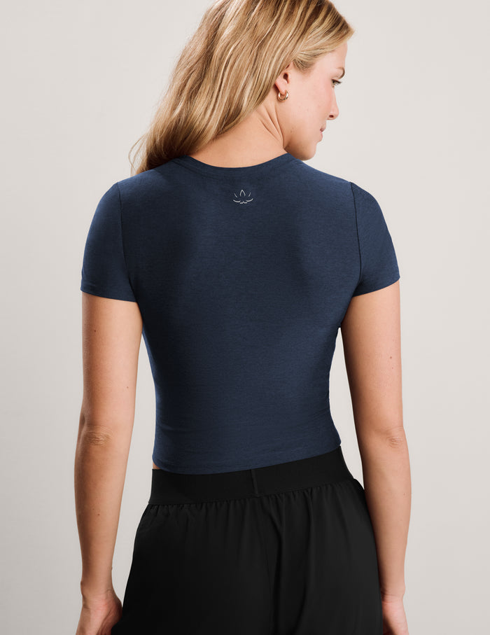 A woman wearing a navy blue short-sleeve crop top and black pants, posing against a light gray studio background.