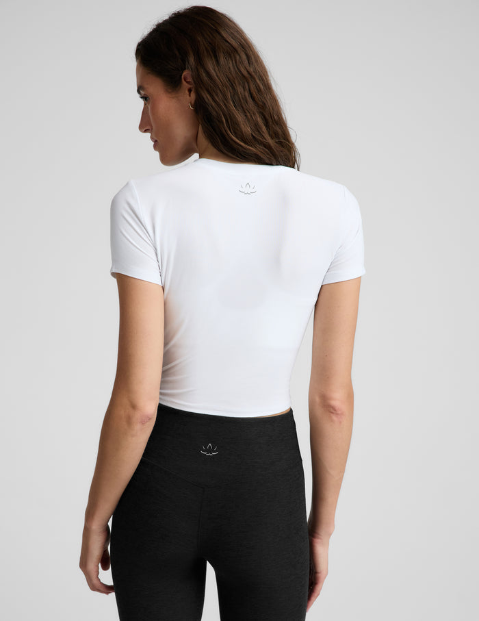 A woman wearing a fitted white short-sleeve crop top and black leggings, photographed against a plain light gray studio background.