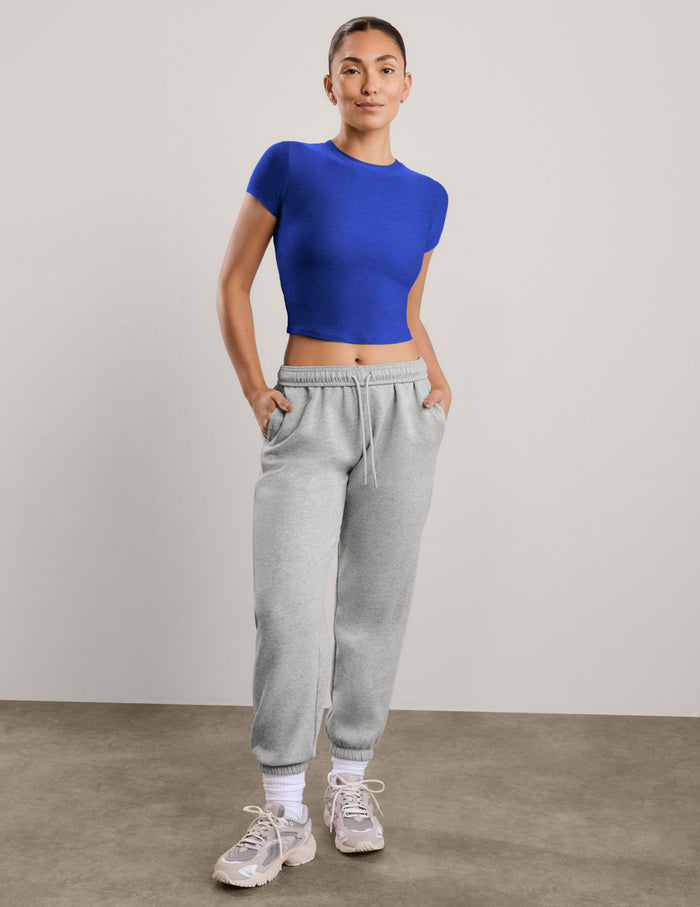 A woman modeling a royal blue short-sleeve crop top paired with light gray sweatpants, standing against a neutral light gray studio background.