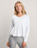 A woman modeling a white long-sleeve crewneck athletic top paired with heather gray leggings, set against a plain light gray studio background.