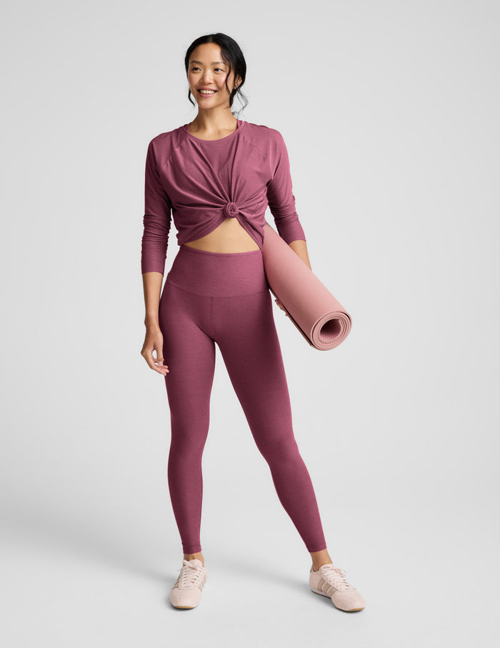 Woman wearing a purple long sleeve top and leggings against a light grey background.