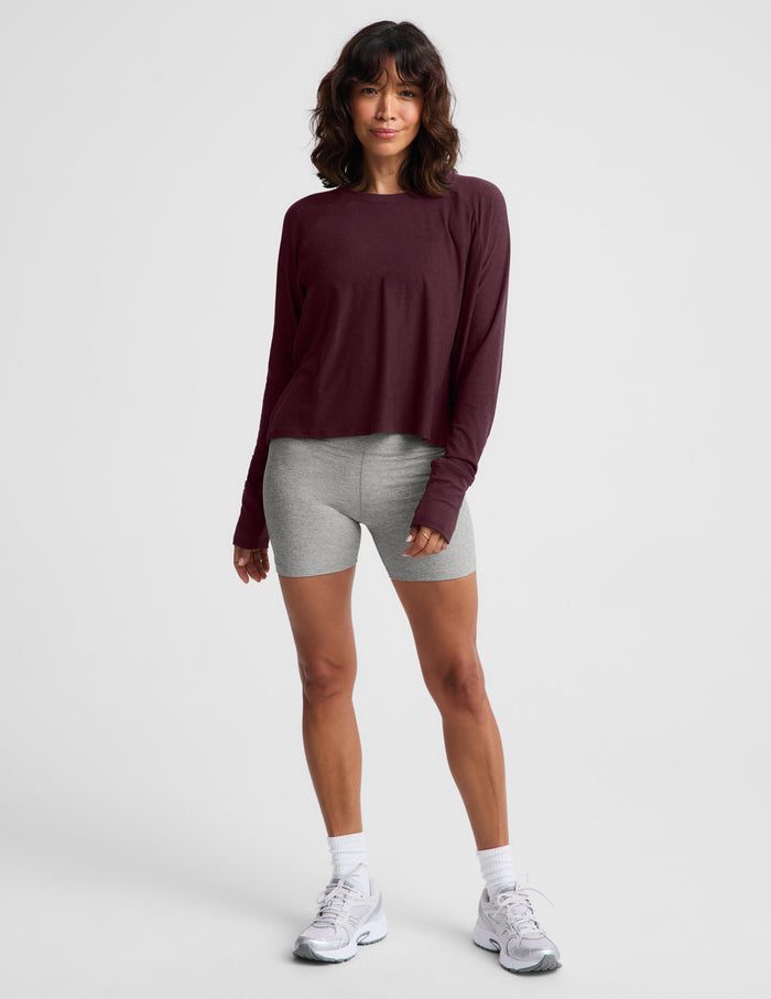 A woman wearing a dark burgundy long-sleeve crewneck top and heather gray bike shorts, standing against a light gray studio background.