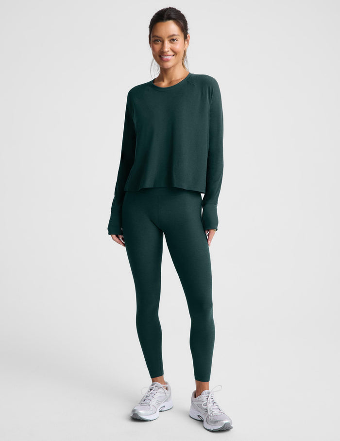 A woman modeling a dark forest green long-sleeve cropped top and matching leggings against a light gray studio background.