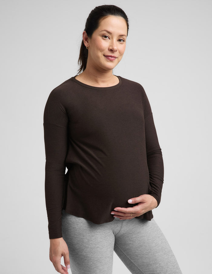 Pregnant woman wearing a brown long sleeve maternity top and gray leggings against a plain background.