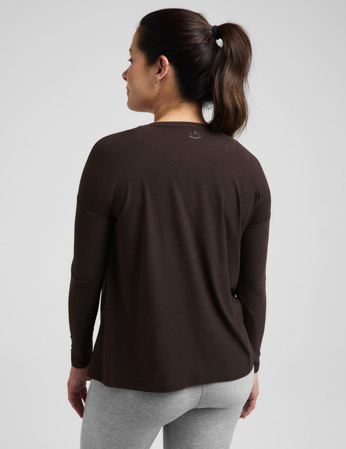 Pregnant woman wearing a brown long sleeve maternity top and gray leggings against a plain background.
