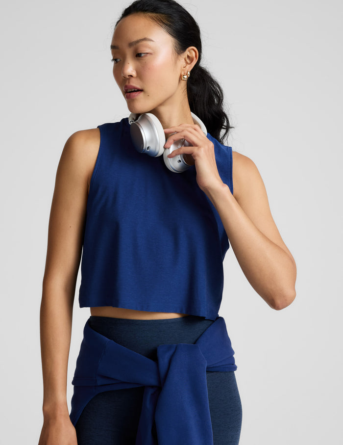 Woman wearing a blue sleeveless top and dark pants holding white headphones against a light gray background.