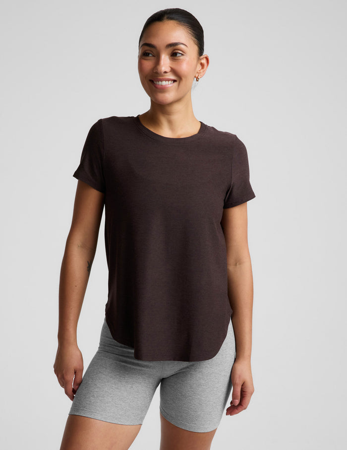 A woman modeling a dark brown short-sleeve crewneck top paired with heather gray bike shorts, photographed against a solid light gray studio background.