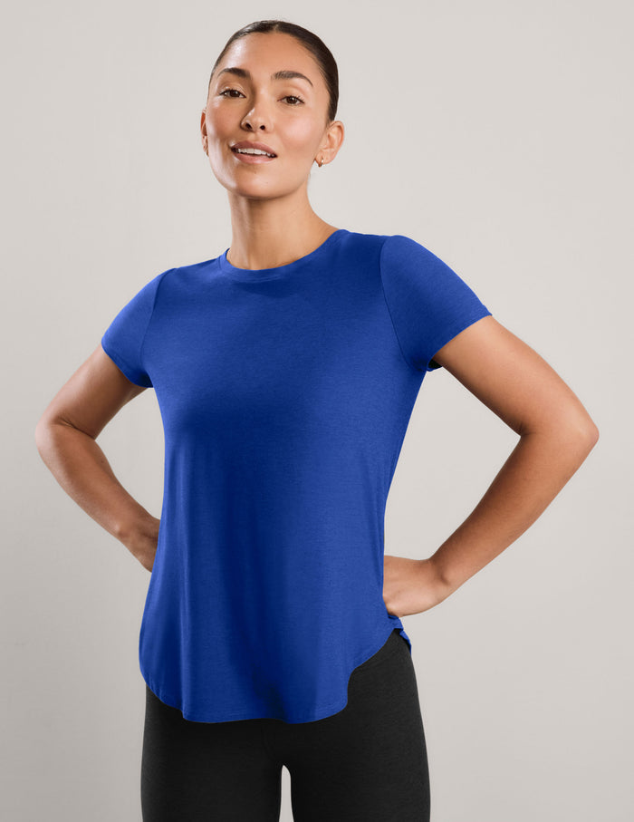 A woman modeling a short-sleeve athletic top in a solid royal blue color, paired with black leggings, against a neutral light gray studio background.