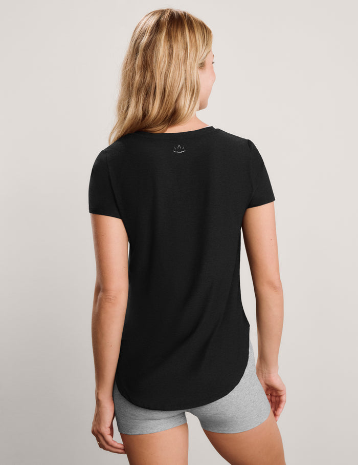 A blonde woman wearing a black short-sleeve crewneck t-shirt and light gray heathered athletic shorts, standing against a neutral light gray studio background.