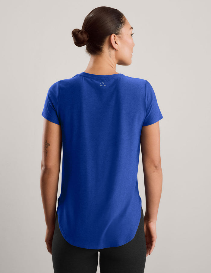 A woman modeling a short-sleeve athletic top in a solid royal blue color, paired with black leggings, against a neutral light gray studio background.