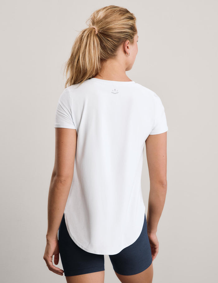 A woman viewed from the back wearing a white short-sleeve athletic top and dark navy fitted shorts against a plain light gray studio background.