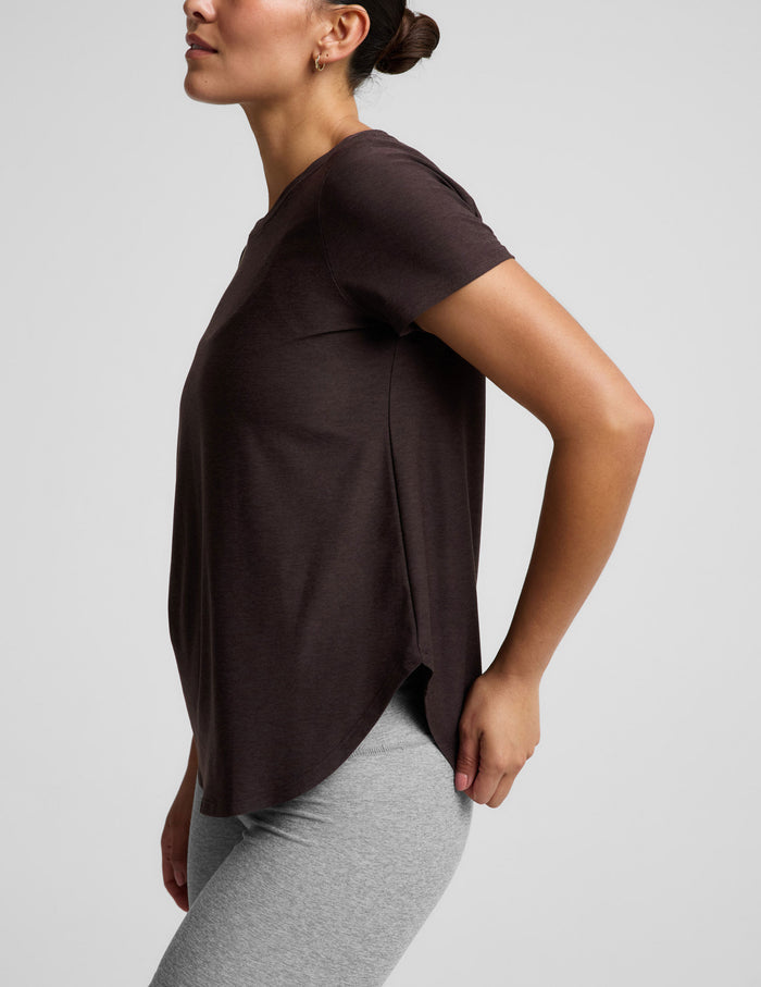 A woman modeling a dark brown short-sleeve crewneck top paired with heather gray bike shorts, photographed against a solid light gray studio background.