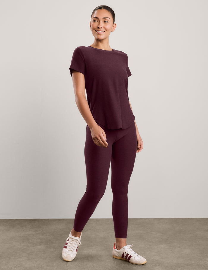 A woman wearing a short sleeve burgundy t-shirt and matching leggings against a plain studio background.