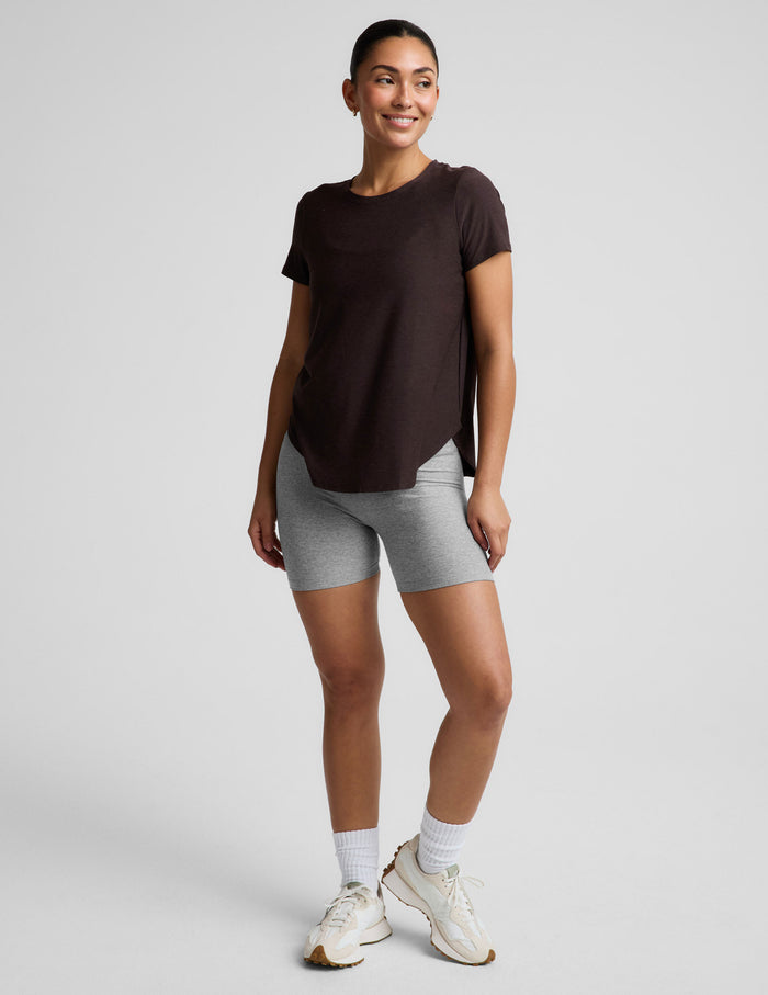 A woman modeling a dark brown short-sleeve crewneck top paired with heather gray bike shorts, photographed against a solid light gray studio background.