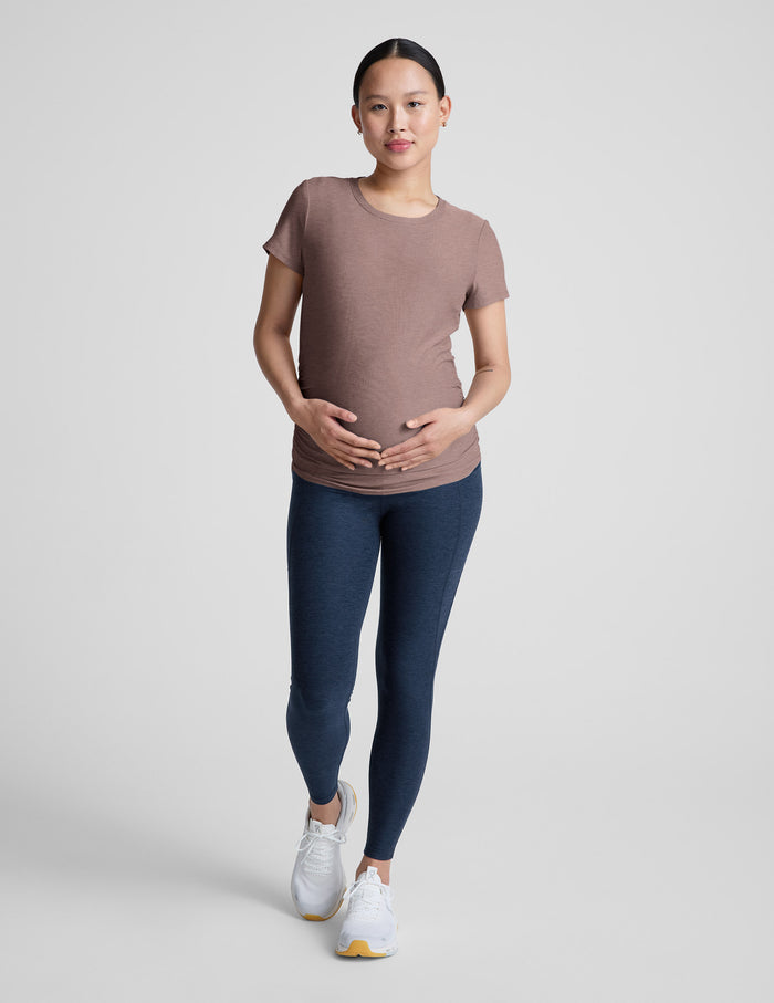 Pregnant woman wearing a brown shirt and blue leggings against a plain background.