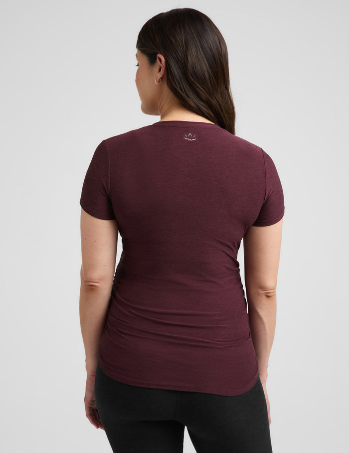 A pregnant woman wearing a short-sleeve crewneck maternity t-shirt in maroon and black bike shorts, standing against a light gray studio background.