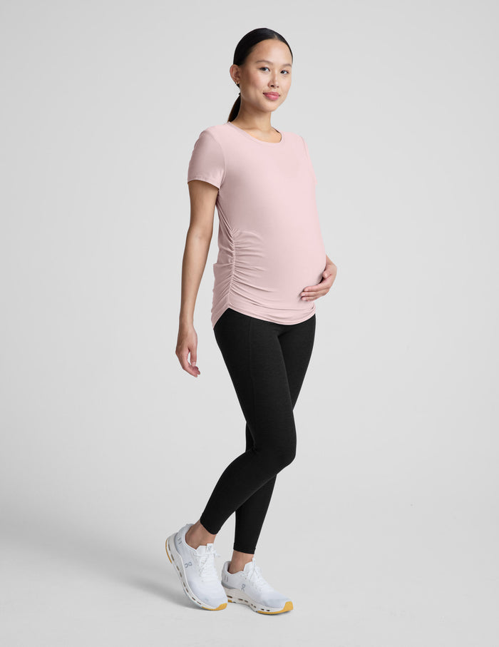Woman wearing a pink maternity t-shirt and black maternity leggings on a plain background.