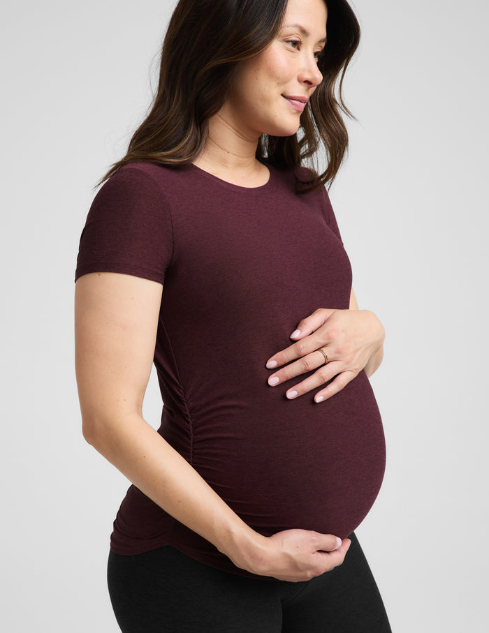 A pregnant woman wearing a short-sleeve crewneck maternity t-shirt in maroon and black bike shorts, standing against a light gray studio background.