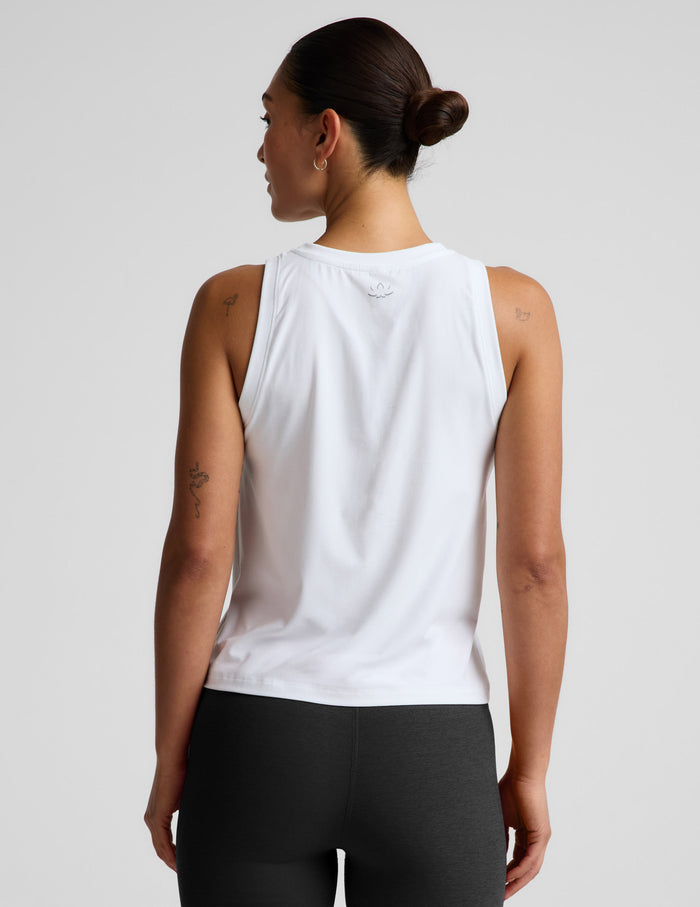 A woman modeling a white sleeveless athletic tank top paired with black biker shorts, photographed against a clean light gray studio background.