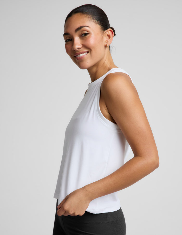 A woman modeling a white sleeveless athletic tank top paired with black biker shorts, photographed against a clean light gray studio background.