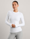 A woman modeling a fitted white long-sleeve crewneck athletic top against a plain light gray studio background.