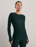 A woman modeling a dark forest green long-sleeve crewneck top and matching pants against a neutral light gray studio background.