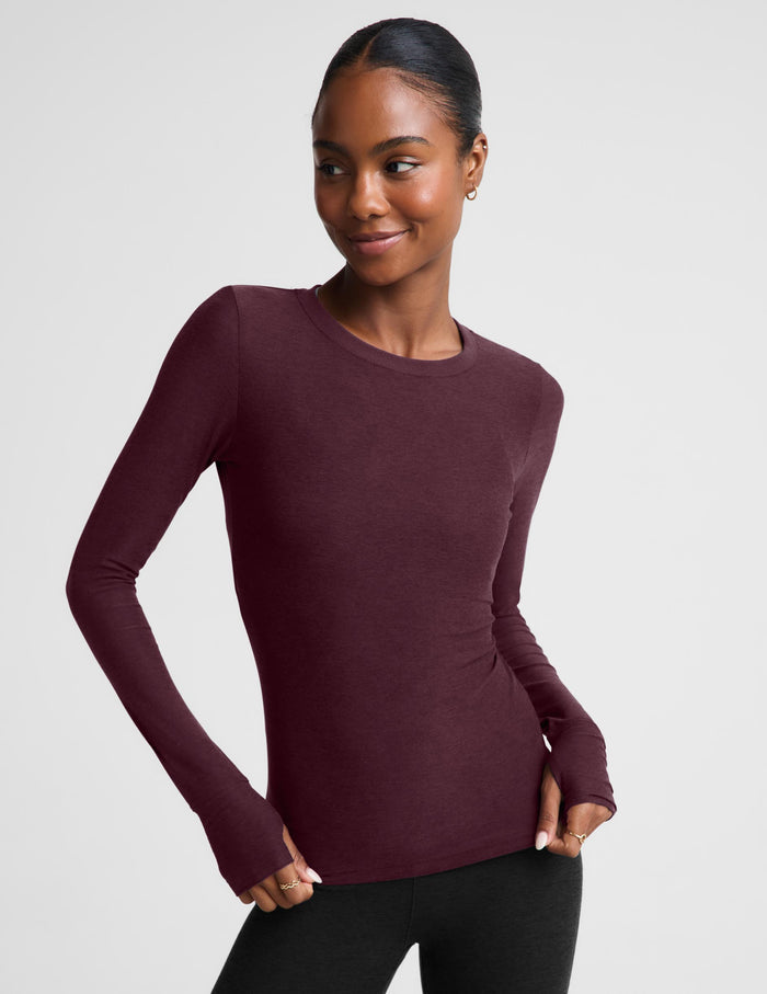 A woman modeling a long-sleeve crewneck top in a deep burgundy color, paired with black pants, against a solid light gray background.
