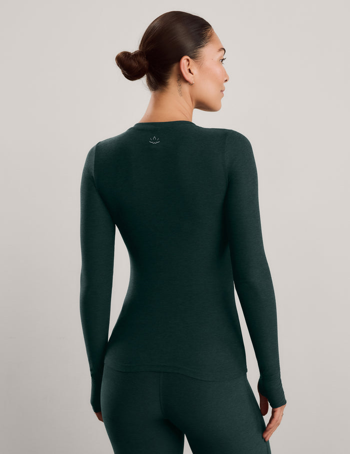 A woman modeling a dark forest green long-sleeve crewneck top and matching pants against a neutral light gray studio background.
