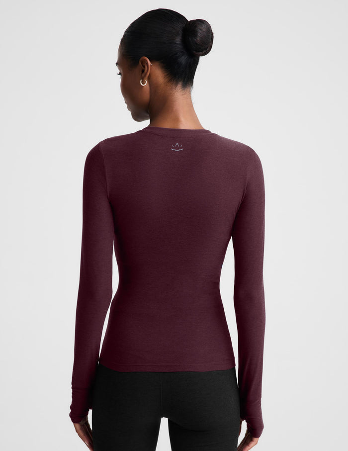 A woman modeling a long-sleeve crewneck top in a deep burgundy color, paired with black pants, against a solid light gray background.
