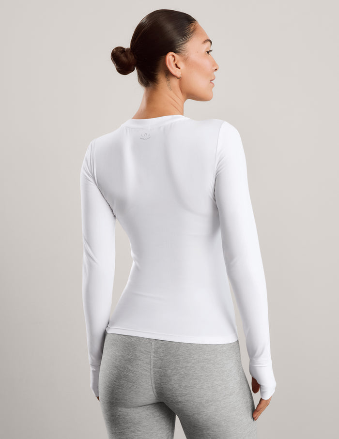 A woman modeling a fitted white long-sleeve crewneck athletic top against a plain light gray studio background.