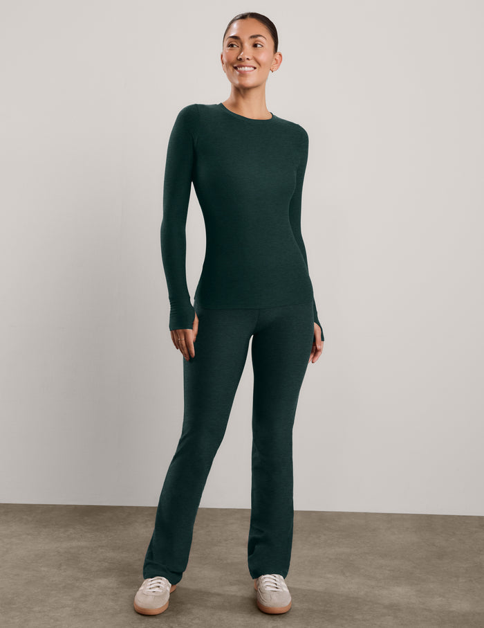 A woman modeling a dark forest green long-sleeve crewneck top and matching pants against a neutral light gray studio background.