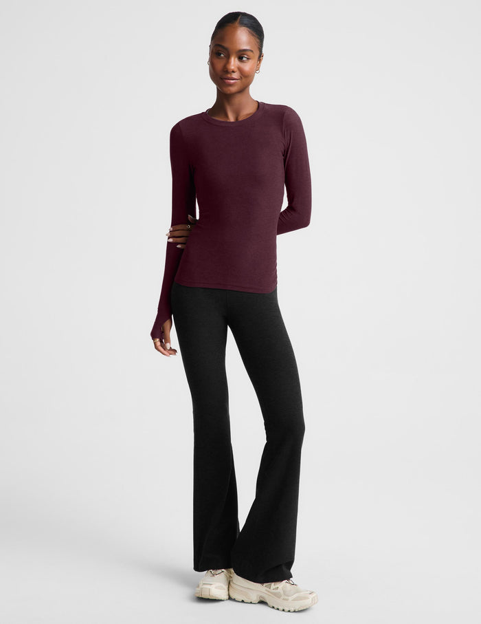 A woman modeling a long-sleeve crewneck top in a deep burgundy color, paired with black pants, against a solid light gray background.