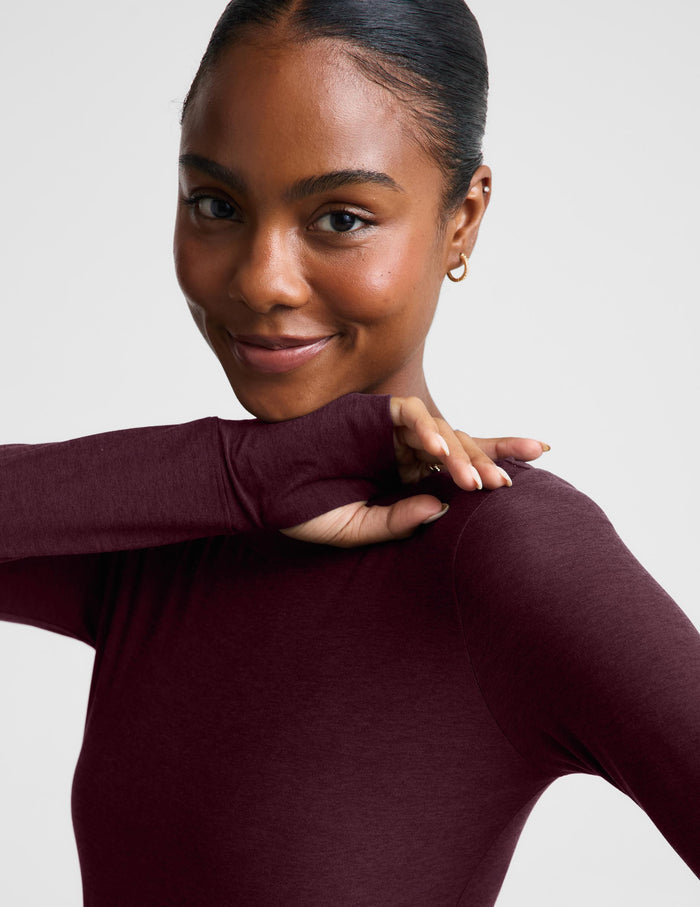 A woman modeling a long-sleeve crewneck top in a deep burgundy color, paired with black pants, against a solid light gray background.