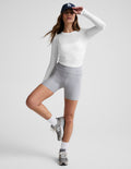 Woman wearing a white long sleeve top with shirring on sides and gray shorts against a plain background.