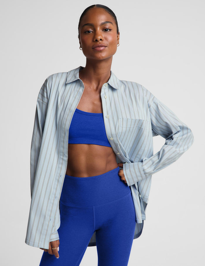 A model wearing a light blue and white vertically striped button-down shirt over a royal blue sports bra and matching royal blue leggings, posed against a solid light gray background.
