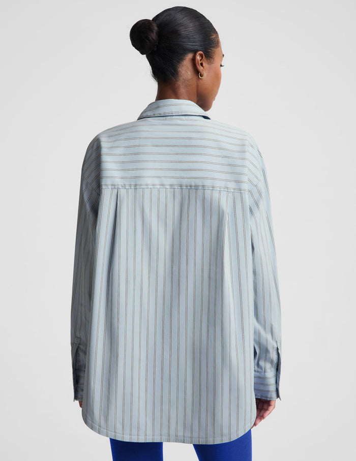 A woman shown from the back wearing an oversized light blue and white vertically striped button-up shirt, paired with blue pants, against a plain light gray studio background.