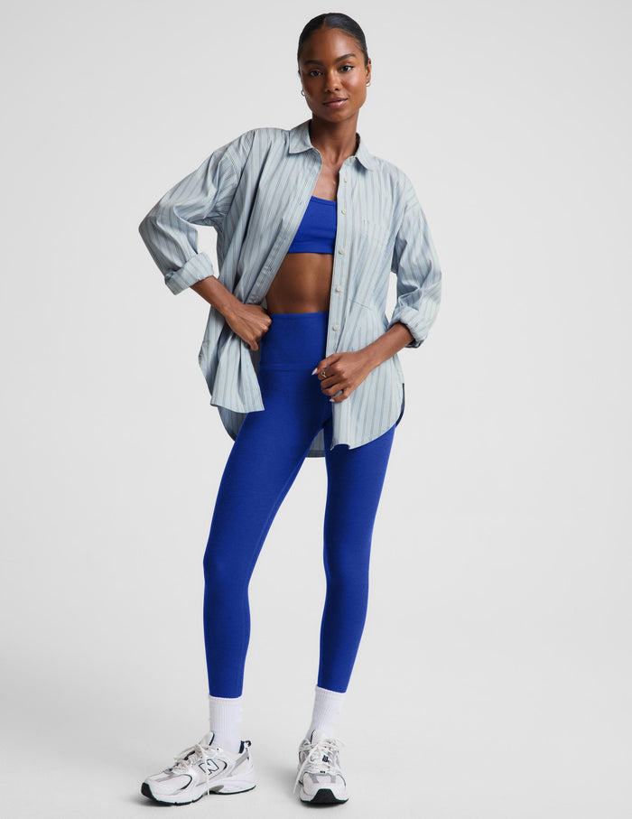 A model stands against a light gray studio background wearing a light blue and white vertically striped button-up shirt over a royal blue sports bra and matching royal blue leggings, with white socks and white sneakers.