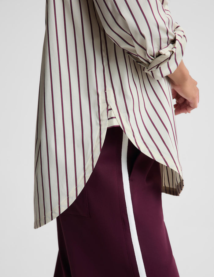 Close-up of a person wearing a cream-colored blouse with thin dark burgundy stripes and dark burgundy satin trousers with a white side stripe, against a light gray studio background.