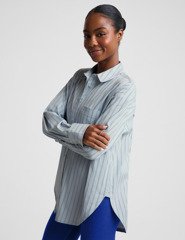 A woman modeling a light blue and white vertically striped button-up shirt with a collar and long sleeves, paired with blue leggings, against a plain light gray studio background.