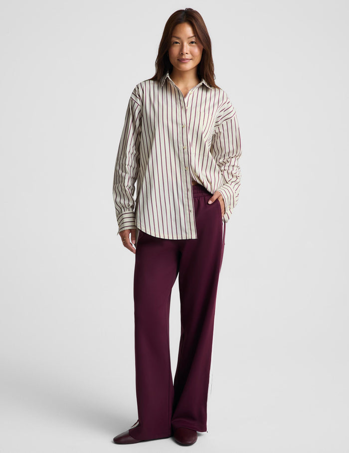 A woman modeling a cream-colored long-sleeve button-up shirt with thin dark vertical stripes, paired with dark plum wide-leg trousers, against a light gray studio background.