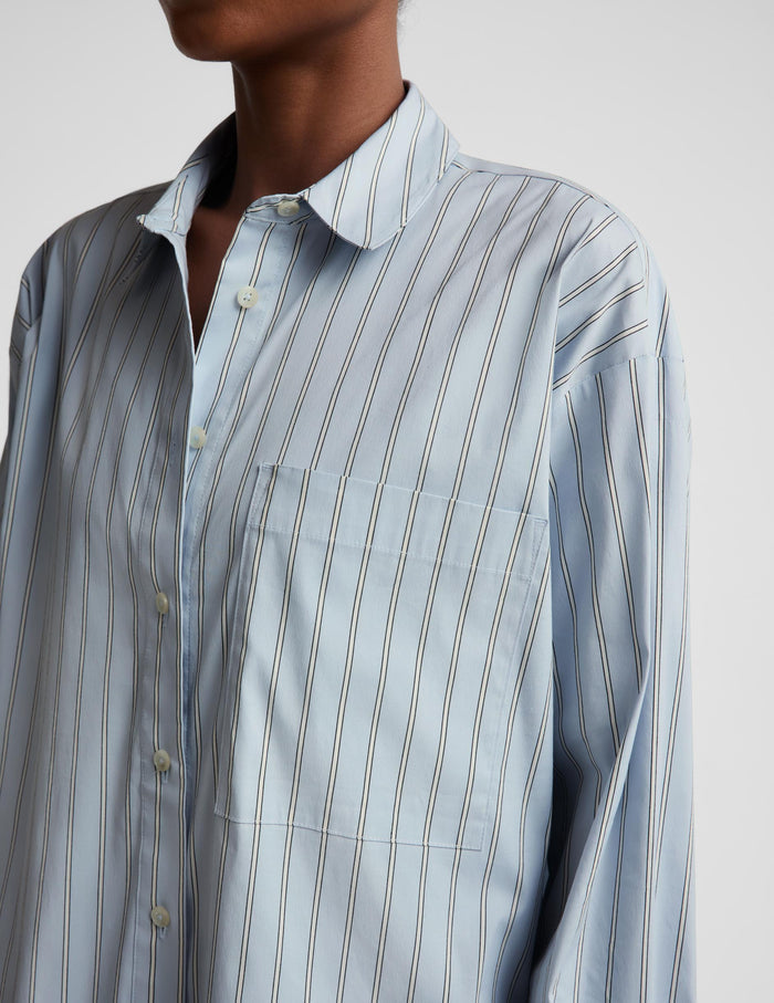 A close-up of a person wearing a light blue button-up shirt with thin vertical dark stripes and a chest pocket, photographed against a plain light background.