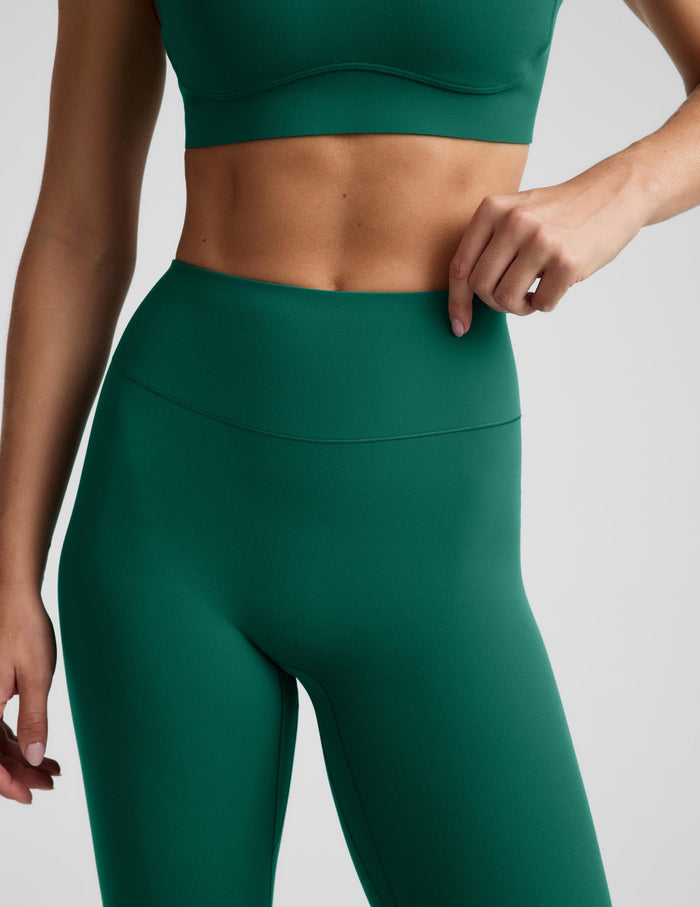 A close-up of a model wearing dark green athletic leggings and a matching sports bra against a light gray studio background.
