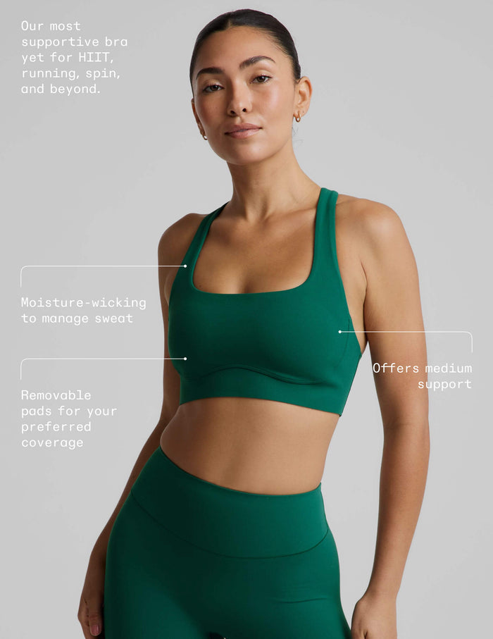 A woman wearing a dark green athletic sports bra and matching high-waisted bottoms against a light gray studio background.