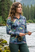 Woman in a patterned jacket holding a fly fishing rod by a river with trees in the background
