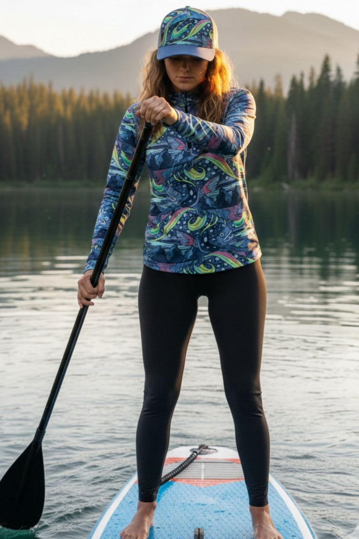 Woman paddleboarding on a lake wearing a colorful patterned shirt and cap.