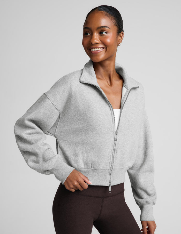 A woman wearing a light gray heather cropped zip-up jacket with balloon sleeves and dark brown leggings, posing against a solid light gray background.