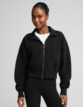A woman wearing a black cropped zip-up sweatshirt with a stand collar over a white top and black leggings, photographed against a plain light gray studio background.