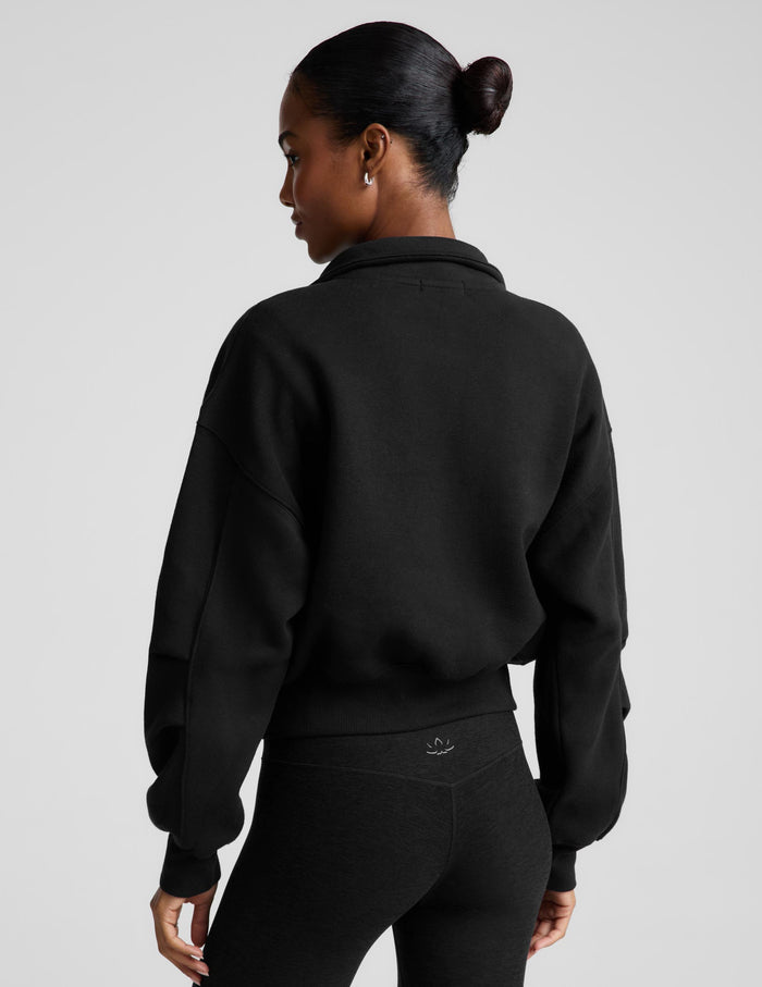 A woman wearing a black cropped zip-up sweatshirt with a stand collar over a white top and black leggings, photographed against a plain light gray studio background.