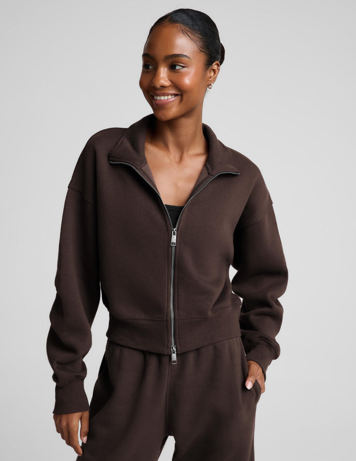A woman wearing a dark brown zip-up sweatshirt and matching sweatpants against a light gray studio background.